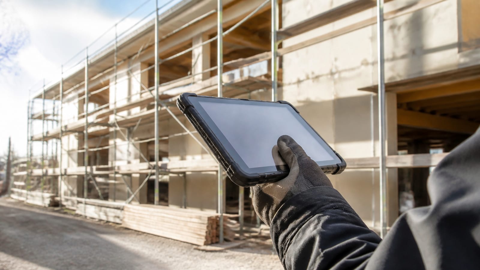 Eine individuelle KI-App entwickeln zu lassen lohnt sich dort, wo draußen gearbeitet wird. Eine behandschuhte Hand hält ein Tablet in Schutzhülle auf der Baustelle, dahinter unscharf ein Gerüst. (KI-generierte Illustration)