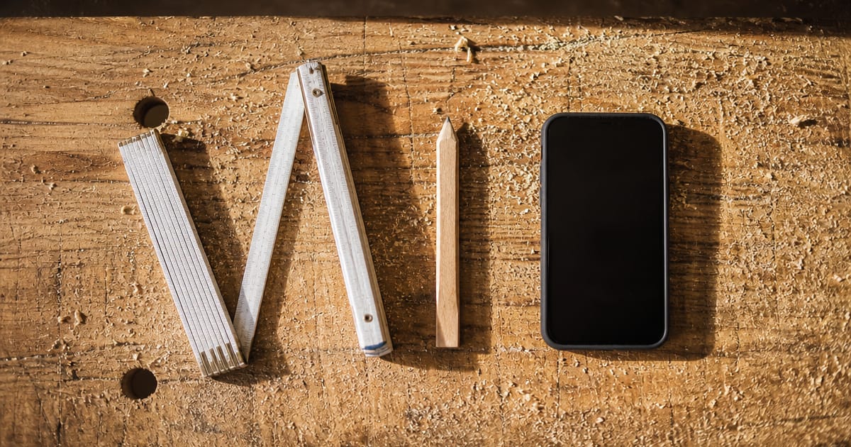 KI-Anwendungsfälle im Handwerk beginnen bei den Aufgaben, die täglich anfallen. Aufsicht auf Zollstock, Zimmermannsbleistift und Smartphone nebeneinander auf einer rauen Holzfläche. (KI-generierte Illustration)